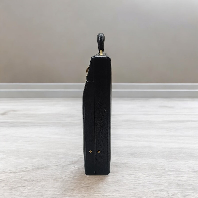 May include: A black rectangular handbag with a small handle. The bag has gold-colored accents and hardware. The side profile shows a sleek, structured design, suggesting a vintage or classic style. The bag is likely made of leather or a similar material.