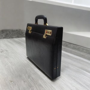 May include: A black briefcase with a textured surface, gold-colored hardware, and a combination lock. The briefcase has a handle and is closed. The briefcase is rectangular and appears to be made of leather.