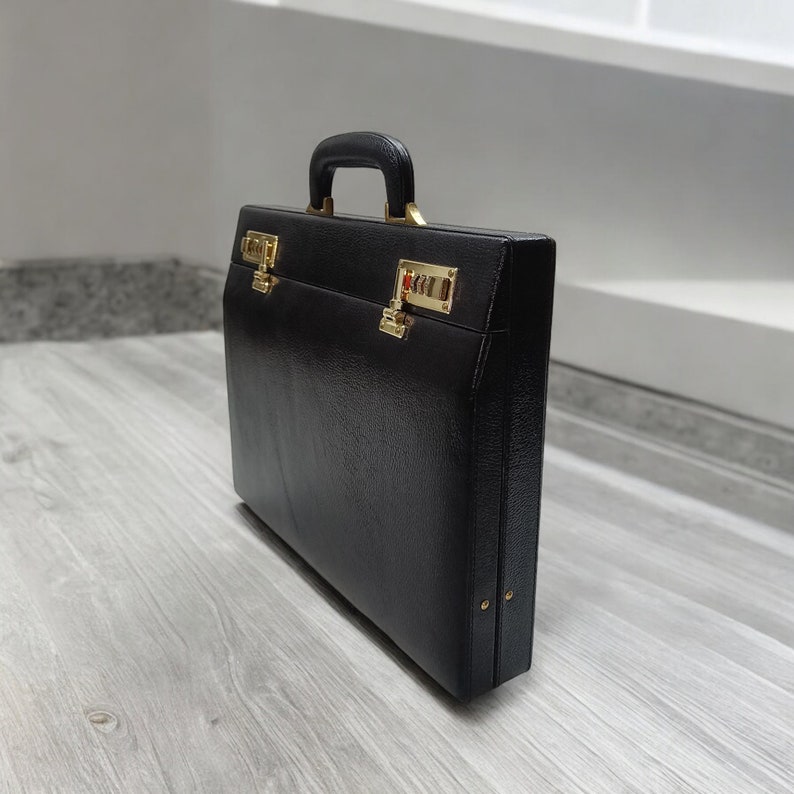 May include: A black briefcase with a textured surface, gold-colored hardware, and a combination lock. The briefcase has a handle and is closed. The briefcase is rectangular and appears to be made of leather.