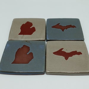 May include: Set of four ceramic coasters featuring the outlines of Michigan and the Upper Peninsula in red on a beige and blue background.