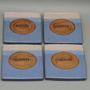 May include: Set of four blue and white ceramic coasters with a brown circle in the center that says "Detroit" in a wood-burned font.