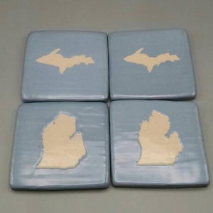 May include: Set of four blue ceramic coasters with white outlines of the Upper Peninsula and the state of Michigan.