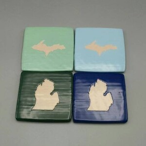 May include: Four square ceramic coasters with a white outline of the Upper Peninsula of Michigan. The coasters are in the colors green, blue, dark green, and dark blue.