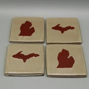 May include: Set of four square ceramic coasters featuring the outline of the state of Michigan in red. Two coasters show the state in its traditional shape, while the other two show the state in its "mitten" shape.