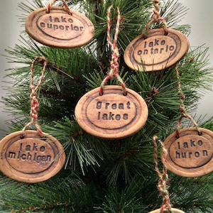 May include: Six brown ceramic ornaments with the names of the Great Lakes written on them. The ornaments are hanging on a small artificial Christmas tree.