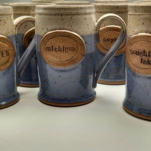 May include: A collection of six ceramic mugs with speckled glaze. Each mug has a different inscription in a brown oval on the front: "up north", "michigan", "boyne", "houghton lake", "traverse city", and "grand rapids".