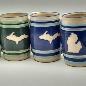 May include: Four ceramic mugs with white outlines of the Upper Peninsula of Michigan on a green, blue, and white striped background.