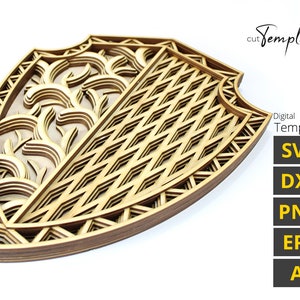 Puede incluir: Un escudo de madera con un diseño en capas, que presenta un patrón geométrico en un lado y un patrón floral en el otro. El escudo está hecho de múltiples capas de madera, creando un efecto 3D. El escudo es una plantilla digital disponible en los formatos SVG, DXF, PNG, EPS y AI.