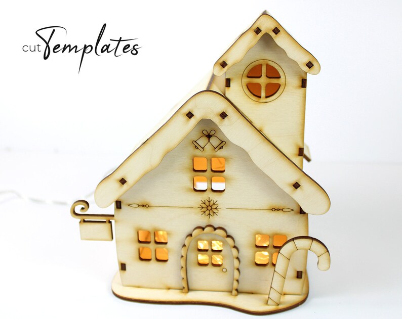 Puede incluir: Una casa de Navidad de madera con un bast&oacute;n de caramelo, una campana y un dise&ntilde;o de copo de nieve. La casa tiene una luz en el interior, lo que la hace brillar.