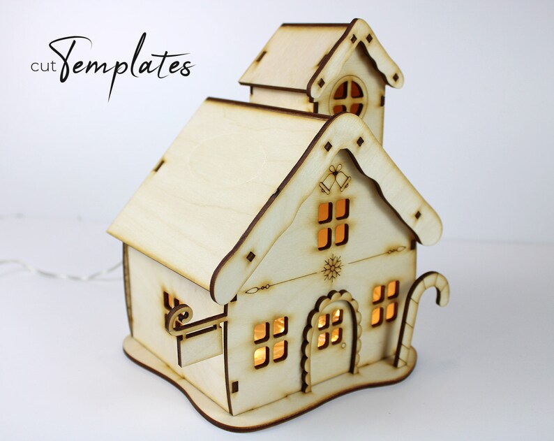 Puede incluir: Una casa de Navidad de madera con un bast&oacute;n de caramelo, campanas y un dise&ntilde;o de copo de nieve. La casa tiene luces en el interior y est&aacute; hecha de madera sin terminar.