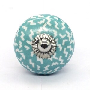 Peut inclure: Un bouton en céramique bleu sarcelle avec un motif géométrique blanc. Le bouton a une base en métal argenté.