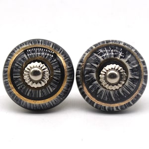 Op de afbeelding: Twee ronde kastknoppen met een zwart-wit patroon, een goudkleurige band en een zilverkleurig midden. De knoppen zijn waarschijnlijk van keramiek of porselein. Ideaal voor meubels.