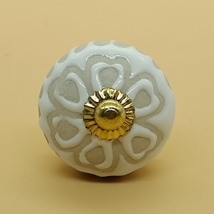 Könnte beinhalten: Ein weißer Keramik-Knopf mit goldener Mitte und weißem Blumenmuster. Der Knopf ist rund und hat eine glatte Oberfläche.