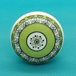 May include: A white ceramic knob with a green and brown floral design. The knob has a circular shape and a raised design.