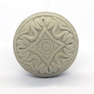 May include: A round, gray, decorative knob with a raised, intricate design. The design features a central diamond shape with a circle inside, surrounded by four stylized leaves.