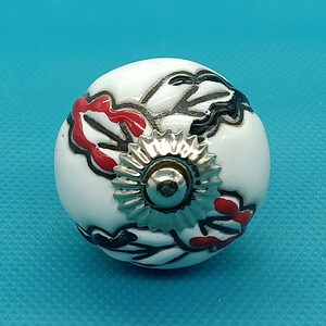 Könnte beinhalten: Weißer Keramik-Knopf mit einem schwarzen und roten Blumendesign und einem silbernen Metallakzent.