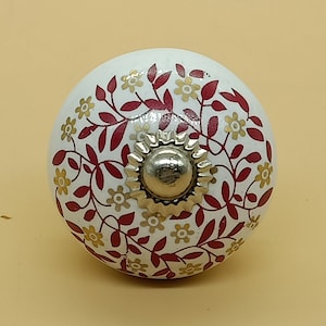Op de afbeelding: Witte keramische knop met een rood en gouden bloemenmotief. De knop heeft een zilveren metalen midden.