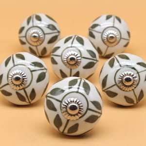 May include: Six white ceramic cabinet knobs with a green leaf design and silver accents.