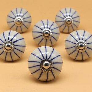 Peut inclure: Six boutons de porte en céramique blanche avec des rayures bleues et des accents argentés. Les boutons ont une forme arrondie, légèrement bombée.
