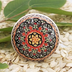 Könnte beinhalten: Ein runder Keramik-Knopf mit einem farbenfrohen Mandala-Design. Das Mandala zeigt komplizierte Muster in Grün-, Orange-, Rot- und Schwarztönen.