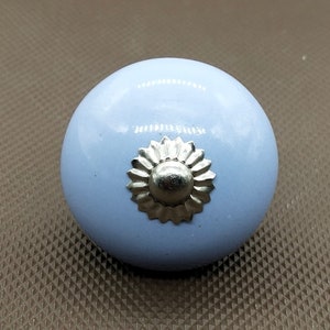 May include: A light blue, round cabinet knob with a silver-colored floral design in the center. The knob has a glossy finish and is designed for furniture. The hardware is a decorative accent for drawers and doors.