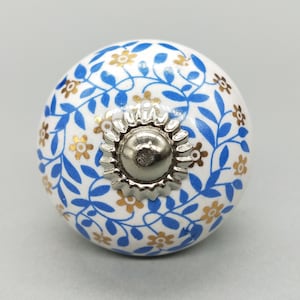 Könnte beinhalten: Weißer Keramik-Knopf mit einem blauen und goldenen Blumenmuster. Der Knopf hat eine silberne Metallmitte.