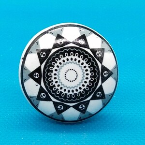 Op de afbeelding: Een witte keramische knop met een zwart-wit geometrisch ontwerp. Het ontwerp heeft een centrale cirkel met een stervormig patroon omgeven door een ring van kleinere cirkels.