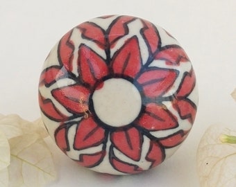May include: A white ceramic knob with a red and black floral design. The knob is round and has a smooth surface.