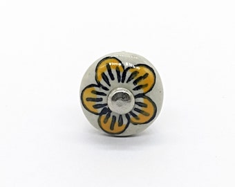 Op de afbeelding: Een witte keramische knop met een geel bloemenontwerp. De bloem heeft zwarte contouren en een zilveren midden.