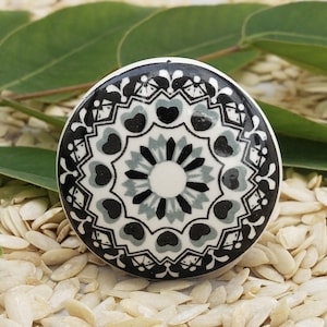 Op de afbeelding: Een ronde, decoratieve kastknop met een witte basis en een gedetailleerd zwart, grijs en wit bloemmotief. De knop is geplaatst tegen een achtergrond van lichtgekleurde, ovale objecten en groene bladeren.