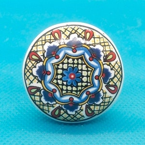 Könnte beinhalten: Ein weißer Keramik-Knopf mit einem farbenfrohen, komplizierten Design. Das Design zeigt ein blaues, gelbes und rotes Blumenmuster mit einer zentralen Sternform.