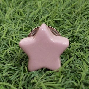 Peut inclure: Une épingle de revers en forme d'étoile rose clair avec un dos argenté. L'épingle est faite d'un matériau brillant et est posée sur un fond d'herbe verte.