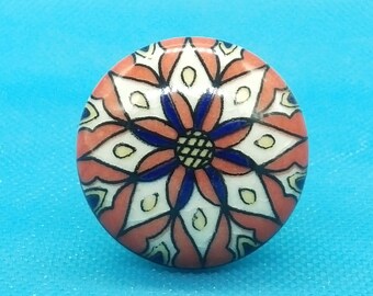 May include: A round ceramic knob with a colorful floral design. The knob is painted in shades of red, white, blue, and yellow. The design features a central flower with intricate details.