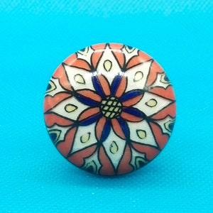 Puede incluir: Un pomo redondo de cerámica con un diseño floral colorido. El pomo está pintado en tonos de rojo, blanco, azul y amarillo. El diseño presenta una flor central con detalles intrincados.