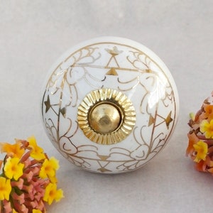 以下が含まれることがあります： 金色の花の模様が施された白いセラミック製のノブ。ノブの中央には金色の金属製の部品があります。