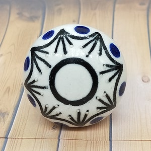 May include: White ceramic knob with a black and blue geometric design. The knob has a circular center with a black outline and a white interior. The design features black radiating lines and blue dots.