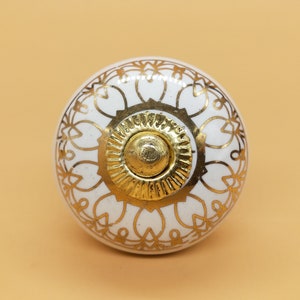May include: White ceramic knob with a gold decorative design. The knob has a round shape and a raised center.