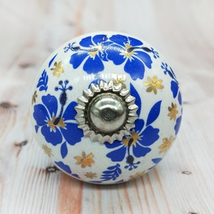 May include: White ceramic knob with a blue and gold floral design. The knob has a silver metal center.
