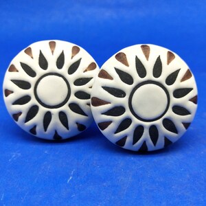 Pode incluir: Dois botões de cerâmica branca com um design de sol preto e marrom. Os botões são redondos e têm um design em relevo.
