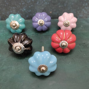 Puede incluir: Seis pomos de armario de cerámica en varios colores, incluyendo rosa, azul, rojo, negro y morado. Cada pomo tiene un diseño en forma de flor con un centro de metal plateado.