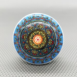 Peut inclure: Un bouton rond en céramique bleu et blanc avec un motif de mandala coloré. Le bouton présente un cercle central avec un motif de fleur rouge et jaune, entouré d'un anneau de feuilles jaunes et vertes et d'un anneau de fleurs rouges et bleues.