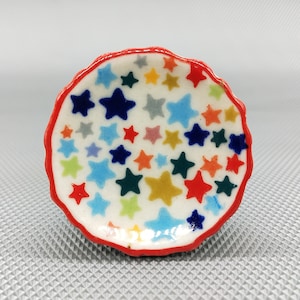 Puede incluir: Un pequeño plato de cerámica blanca con un borde rojo. El plato está decorado con un patrón colorido de estrellas en tonos de azul, rojo, amarillo y verde.