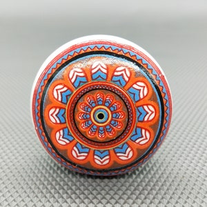 May include: A round, white ceramic knob with a colorful mandala design. The mandala features orange, blue, and white colors in a geometric pattern.