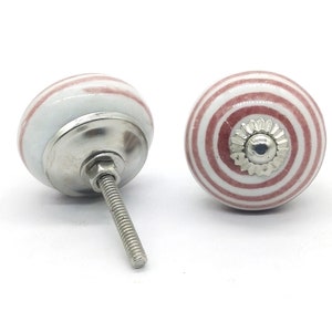 以下が含まれることがあります： 赤色のストライプが入った白いセラミック製のキャビネットノブが2つ。ノブには銀色のネジと銀色の装飾的な部品が中央にあります。