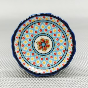 Könnte beinhalten: Ein kleiner, blauer und weißer Keramikteller mit einem roten und gelben Blumendesign in der Mitte. Der Teller hat einen dekorativen Rand mit einem sich wiederholenden Muster aus Blau, Gelb und Rot.