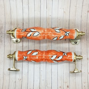 Può includere: Due tiretti per cassetti in ceramica arancione con motivi floreali bianchi e dettagli in ottone. I tiretti sono su uno sfondo in legno.