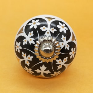 Puede incluir: Pomo de cerámica blanco con un diseño floral negro y dorado. El pomo tiene un centro de metal plateado.