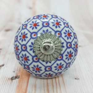 Pode incluir: Um botão de cerâmica branco com um padrão geométrico azul e vermelho e um detalhe em metal prateado. O botão é redondo e tem uma superfície lisa.