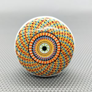 Könnte beinhalten: Ein runder Keramik-Knopf mit einem farbenfrohen Mandala-Design. Das Design zeigt konzentrische Kreise in Gelb, Orange, Rot, Blau und Schwarz. Die Mitte des Mandalas hat einen grünen und weißen Kreis.