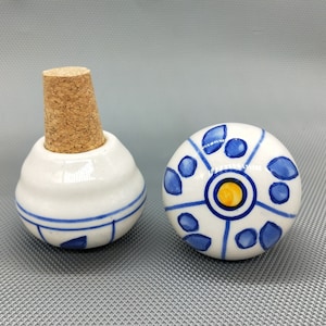 May include: Two ceramic bottle stoppers with blue and white designs. One stopper has a cork top and the other has a round top with a yellow circle in the center.